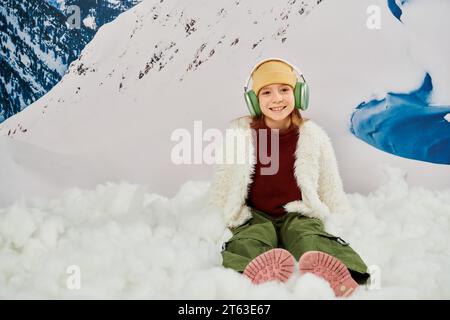 felice ragazza preadolescente in un elegante abbigliamento con cuffie seduto sulla neve e sorridente davanti alla fotocamera, moda Foto Stock