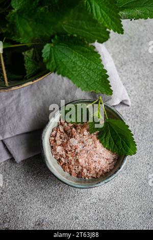 Dall'alto del concetto di cucina vegana, con foglie di ortica fresche in un colino di metallo, accanto a una ciotola di sale rosa himalayano e olio su un dorso grigio Foto Stock