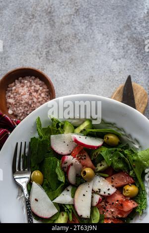 Insalata di verdure con foglie di spinaci biologici freschi, pomodori a fette, aromi e olive presentate in una ciotola bianca accanto a un panno a quadri di legno Foto Stock