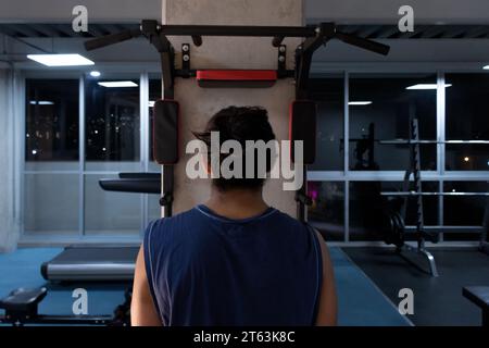 Anonymous Person è in piedi con la schiena alla telecamera, davanti alle attrezzature della palestra e le luci notturne della città brillano delicatamente attraverso le finestre Foto Stock