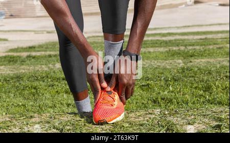Primo piano di mani anonime che legano i lacci delle sue vivaci scarpe da running arancioni su un campo erboso con uno sfondo in cemento Foto Stock