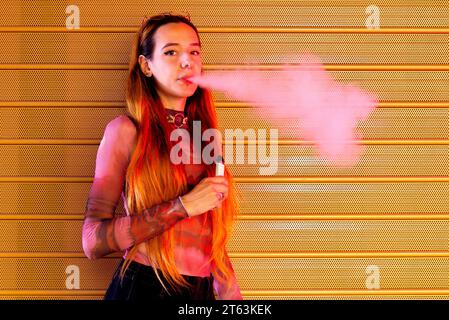 Vista laterale di una giovane donna con i capelli rossi in piedi su uno sfondo arancione perforato, che tiene una sigaretta elettronica ed espira una nuvola di vapore mentre lo Foto Stock