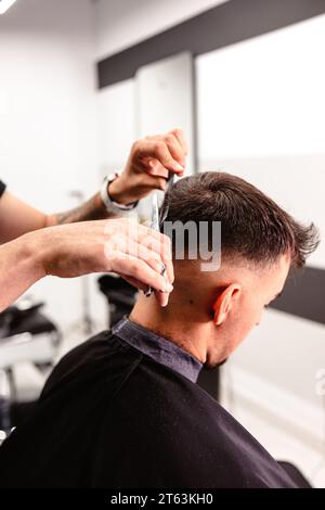 Primo piano delle mani di un barbiere utilizzando sapientemente forbici e pettine per rifinire i capelli di un uomo in un salone moderno. Foto Stock