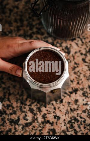 Vista dall'alto del caffè appena macinato ripieno fino all'orlo della macchina per espresso con piano cottura in acciaio inox con un giovane anonimo che tiene la base e. Foto Stock