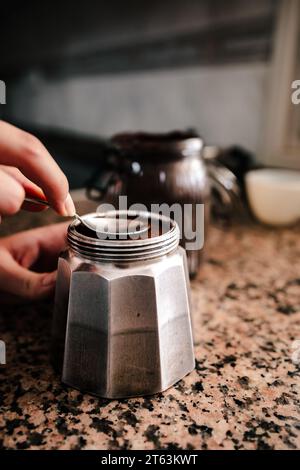Primo piano della mano di un giovane anonimo utilizzando un cucchiaio per aprire o sigillare una macchina per espresso in acciaio inox, in un ambiente da cucina casalingo W. Foto Stock