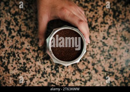 Vista dall'alto del caffè appena macinato ripieno fino all'orlo della macchina per espresso con piano cottura in acciaio inox con un giovane anonimo che tiene la base e. Foto Stock