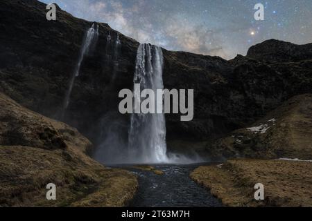 Una maestosa cattura di alte cascate che scendono lungo una scogliera rocciosa scura, con un fiume sereno alla base, il tutto posto sotto un cielo stellato notturno in Islanda Foto Stock