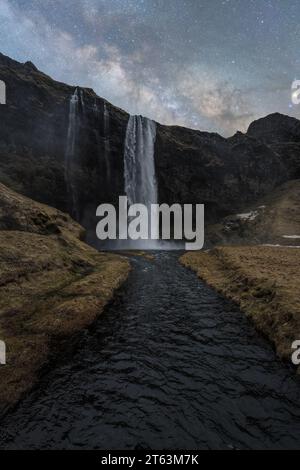 Una maestosa cattura di alte cascate che scendono lungo una scogliera rocciosa scura, con un fiume sereno alla base, il tutto posto sotto un cielo stellato notturno in Islanda Foto Stock