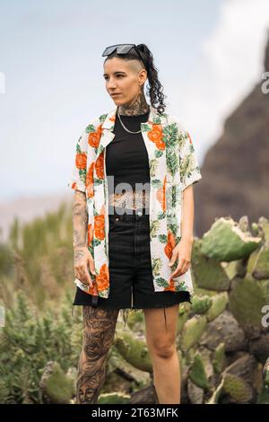 Giovane donna tatuata moderna in abiti casual che guardano lontano mentre si trova in piedi contro la natura sfocata a Masca Tenerife, nelle Isole Canarie Foto Stock