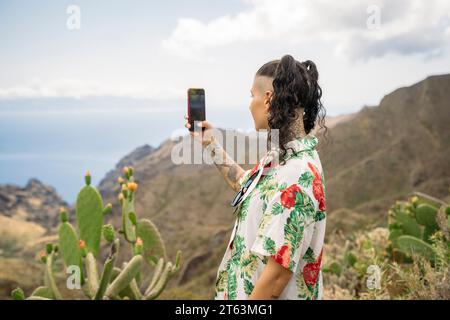 Vista laterale di una giovane donna tatuata in abiti casual che scatta selfie con lo smartphone mentre si trova sullo sfondo delle montagne e del cielo nuvoloso di Masca te Foto Stock