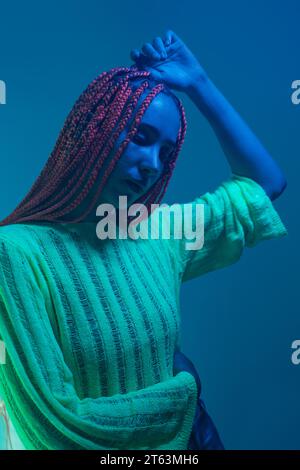 Depressa giovane donna afroamericana con trecce arancioni che contemplano la possibilità di stare in piedi contro la luce brillante al neon blu Foto Stock