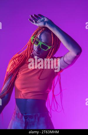 Ritratto di una giovane donna senza emozioni con trecce arancioni che indossa occhiali da sole e posa guardando in basso con sicurezza contro le luci al neon Foto Stock