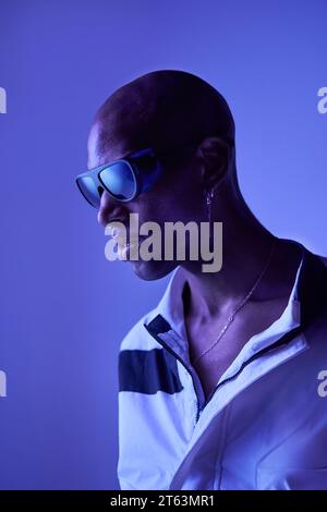 Vista laterale di un giovane uomo calvo, elegante e sicuro, con outfit casual e occhiali da sole che guardano lontano e posano su sfondo blu Foto Stock