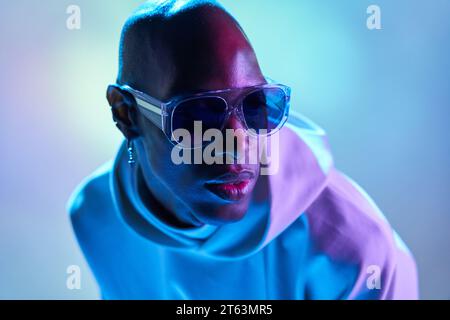Uomo calvo afroamericano serio che indossa orecchini e occhiali da sole che guarda lontano in uno studio moderno con luci illuminanti Foto Stock