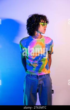 Un giovane uomo dai capelli ricci con una vivace camicia tie-dye si erge su un morbido sfondo blu, indossando occhiali da sole verdi Foto Stock