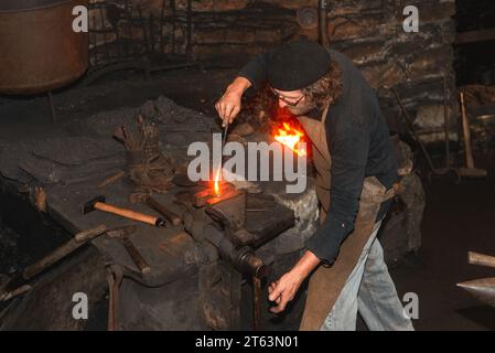 Dall'alto di abile fabbro rivestito di usura tradizionale, forma meticolosamente un raro martello idraulico del XVIII secolo su un'incudine che illustra un'età Foto Stock