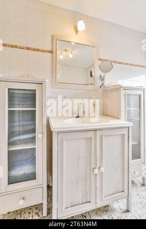 Lampada illuminata e specchio sul lavandino vintage su un armadietto in legno in una cucina rustica con pareti bianche a casa Foto Stock