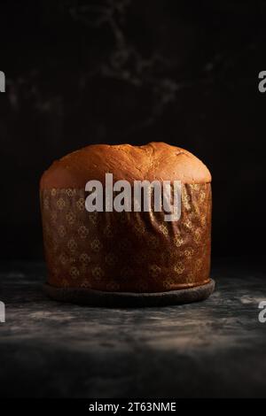 Un panettone appena sfornato, con una ricca crosta dorata, si trova su uno sfondo scuro e lungoso, sottolineandone il calore e la consistenza Foto Stock