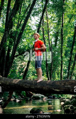 Un viaggiatore felice, con un cappello bianco e delle cuffie sul collo, si trova in cima a un tronco caduto, guardando in alto in una foresta lussureggiante. Foto Stock