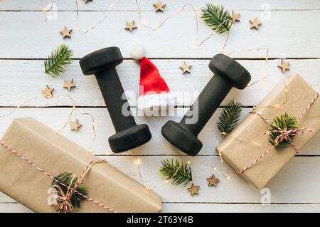 Stile di vita sano durante il periodo di festa di Natale concetto. Manubri neri con ornamenti natalizi su sfondo bianco in legno. Foto Stock