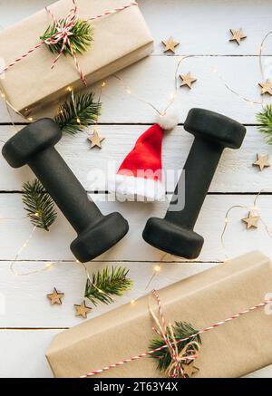 Stile di vita sano durante il periodo di festa di Natale concetto. Manubri neri con ornamenti natalizi su sfondo bianco in legno. Foto Stock