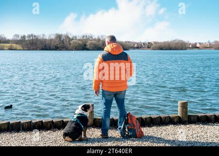 Un pensionato felice con un mantello arancione e uno zaino con un bulldog inglese sulla riva del lago, andando a fare una passeggiata nel giorno primaverile dei vermi. Addestramento dei cani. Tempo libero a reti Foto Stock