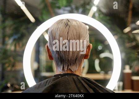 Vista posteriore del giovane uomo con i capelli sbiancati rivolti verso l'anello luminoso nel salone di bellezza e che scatta foto Foto Stock