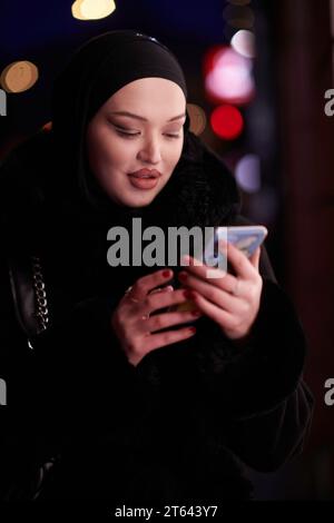 Donna d'affari musulmana Hijabi che controlla il suo telefono sulla strada urbana di notte Foto Stock