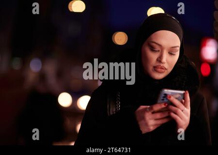 Donna d'affari musulmana Hijabi che controlla il suo telefono sulla strada urbana di notte Foto Stock