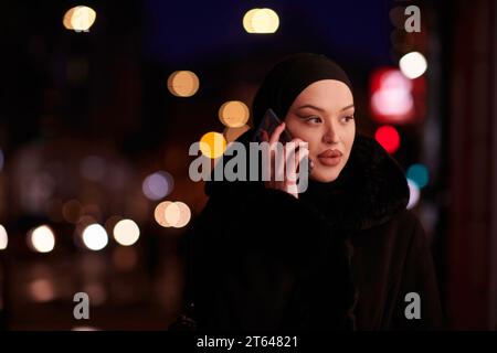 Donna d'affari musulmana Hijabi che controlla il suo telefono sulla strada urbana di notte Foto Stock