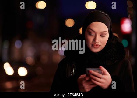 Donna d'affari musulmana Hijabi che controlla il suo telefono sulla strada urbana di notte Foto Stock