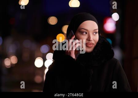 Donna d'affari musulmana Hijabi che controlla il suo telefono sulla strada urbana di notte Foto Stock