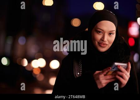 Donna d'affari musulmana Hijabi che controlla il suo telefono sulla strada urbana di notte Foto Stock