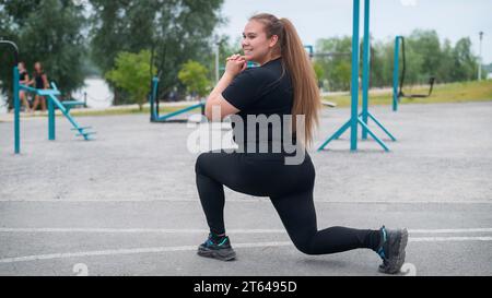 Una bella ragazza grassa con una tuta nera è impegnata nel fitness sul campo sportivo. Una giovane donna si apre all'aperto in una calda giornata estiva. In salute Foto Stock