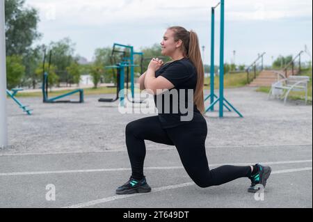 Una bella ragazza grassa con una tuta nera è impegnata nel fitness sul campo sportivo. Una giovane donna si apre all'aperto in una calda giornata estiva. In salute Foto Stock