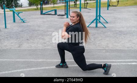 Una bella ragazza grassa con una tuta nera è impegnata nel fitness sul campo sportivo. Una giovane donna si apre all'aperto in una calda giornata estiva. In salute Foto Stock