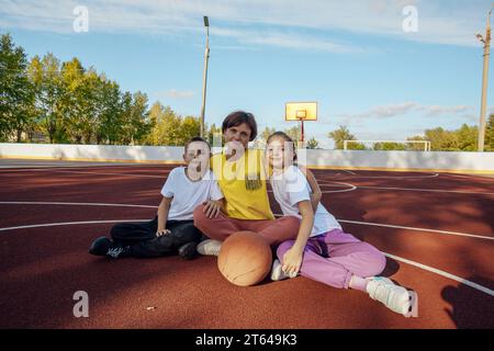Abbracci all'aperto: La madre e i bambini adolescenti si coccolano delicatamente al sole sul campo da basket Foto Stock