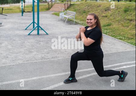 Una bella ragazza grassa con una tuta nera è impegnata nel fitness sul campo sportivo. Una giovane donna si apre all'aperto in una calda giornata estiva. In salute Foto Stock