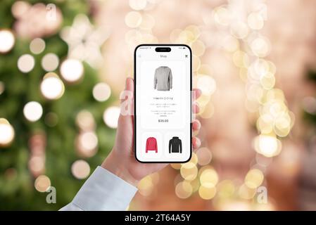 La mano di una donna tiene un telefono cellulare con una moderna app di e-commerce, impegnata nello shopping natalizio. Il bokeh natalizio delle luci dell'albero di Natale crea un holida Foto Stock