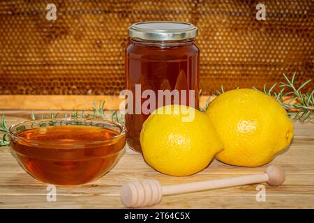 Vaso di miele, dispenser, recipiente con miele, rametto di rosmarino e due limoni su legno con un vero favo sullo sfondo Foto Stock