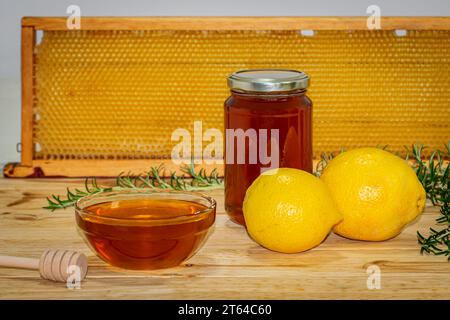 Vaso di miele, dispenser, recipiente con miele, rametto di rosmarino e due limoni su legno con un vero favo sullo sfondo Foto Stock