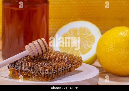 Primo piano di un pezzo di favo e di un dispenser su un piatto di legno, con un barattolo di miele, un limone e mezzo e un vero favo sullo sfondo Foto Stock