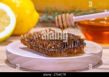 Primo piano di un dispenser che gocciola il miele su un pezzo di favo posto su un piatto di legno, con una ciotola di miele, un rametto di rosmarino, un limone e mezzo Foto Stock