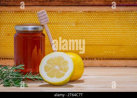 Vaso di miele con dispenser supportato, un rametto di rosmarino e un limone e mezzo su legno con un vero favo sullo sfondo. Spazio per il testo Foto Stock