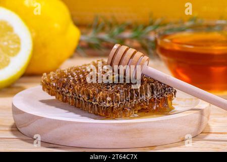 Primo piano di un pezzo di favo e di un dispenser su un piatto di legno, con una ciotola di miele, un rametto di rosmarino, un limone e mezzo e un vero favo Foto Stock