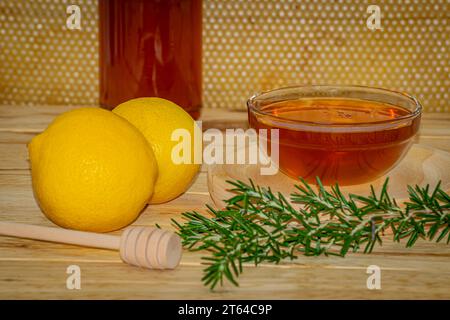 Vaso del miele, dispenser, una ciotola con miele su un piatto di legno, un rametto di rosmarino e due limoni su legno con un vero favo sullo sfondo Foto Stock