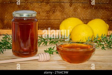 Vaso di miele, dispenser, recipiente con miele, rametto di rosmarino e due limoni su legno con un vero favo sullo sfondo Foto Stock