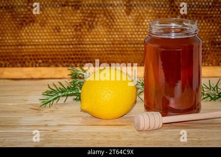 Vaso di miele senza coperchio, dispenser, rametto di rosmarino e limone su legno con un vero favo a nido d'ape sullo sfondo Foto Stock