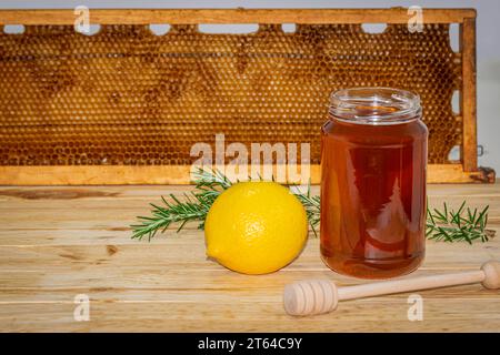 Vaso di miele senza coperchio, dispenser, rametto di rosmarino e limone su legno con un vero favo a nido d'ape sullo sfondo. Spazio per il testo Foto Stock