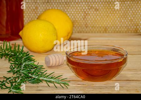 Vaso di miele, dispenser, recipiente con miele, rametto di rosmarino e due limoni su legno con un vero favo sullo sfondo Foto Stock